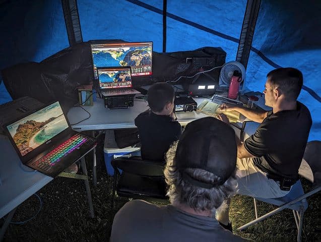 Amateur radio operators working at a Field Day event in a tent with multiple monitors and radios.